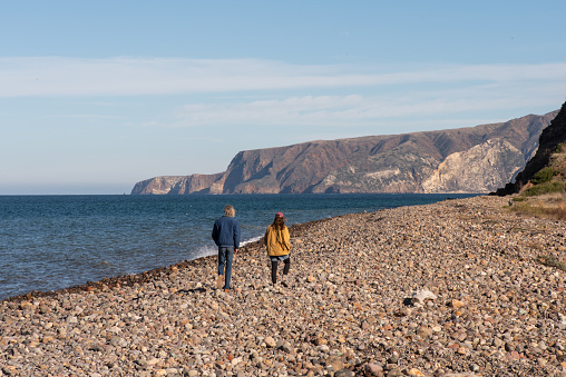 A man and a woman walk down a rocky beach in the Channel Islands, California while waiting for their ferry to arrive.