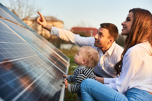 Man shows his family the solar panels on the plot near the house during a warm day. Young woman with a kid and a man in the sun rays look at the solar panels.