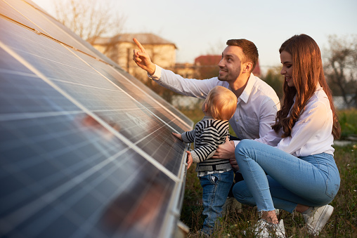 Side view shot of a young modern family with a little baby boy getting acquainted with solar panel on a sunny day, green alternative energy concept