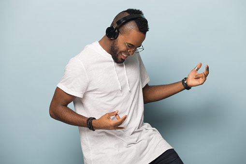 Overjoyed african American millennial man in modern earphones isolated on blue studio background have fun play imaginary guitar, excited biracial man in headphones listen to music dancing