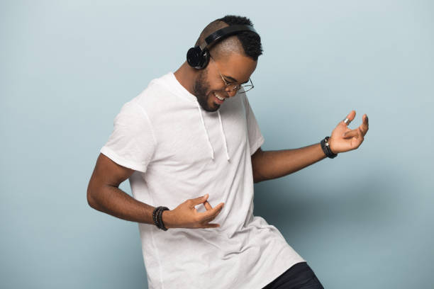 Overjoyed biracial man in earphones have fun dancing Overjoyed african American millennial man in modern earphones isolated on blue studio background have fun play imaginary guitar, excited biracial man in headphones listen to music dancing happy people audio stock pictures, royalty-free photos & images