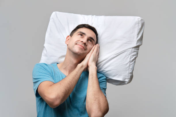 istock Tired brunet man in a blue tee resting on a white pillow isolated over grey background. 1198592206