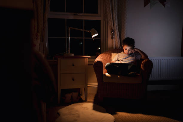 Relaxing in the Snug Bedroom A young boy using a digital tablet while sitting on a chair. He is concentrating while playing on the tablet in a low lit room. low lighting stock pictures, royalty-free photos & images