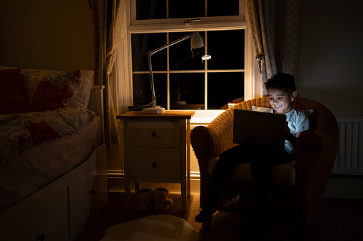 A wide shot of a boy using a digital tablet while sitting on a chair. He is laughing while playing on the tablet in a low lit room.