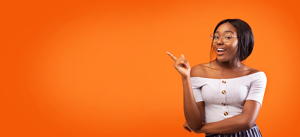 African American Girl Pointing Finger At Copy Space Smiling And Posing Over Orange Studio Background. Special Offer Concept. Panorama