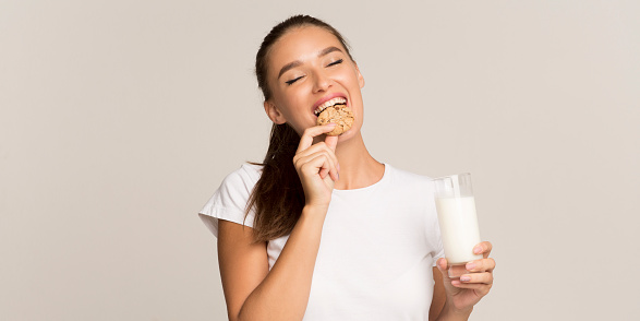 Millennial Girl Drinking Glass Of Milk And Eating Cookies Standing Over Gray Studio Background. Panorama