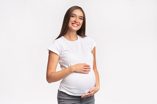 Happy Pregnancy Concept. Expectant Mother-To-Be Touching Belly Smiling At Camera Standing Over White Background. Studio Shot
