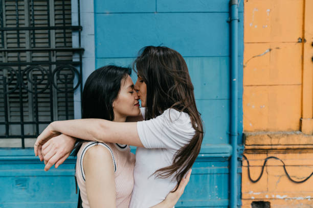 молодая взрослая пара обнимает и целуется против синей и оранжевой стены - lesbian homosexual kissing homosexual couple стоковые фото и изображения