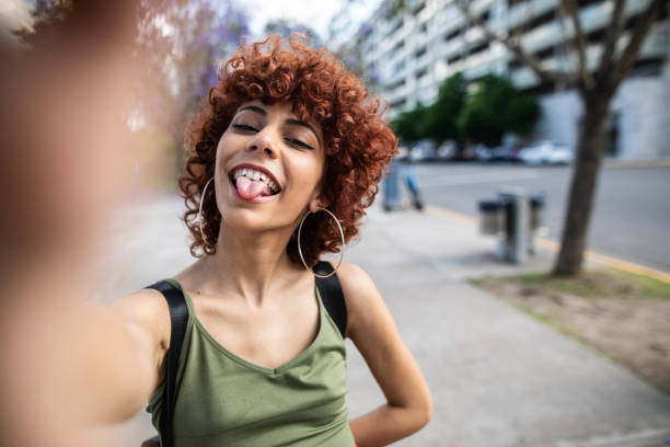 Mischievous Latin girl taking selfie outdoor Portrait of young Latin woman with curly red hair , sticking out tongue and holding camera while taking selfie dyed red hair photos stock pictures, royalty-free photos & images