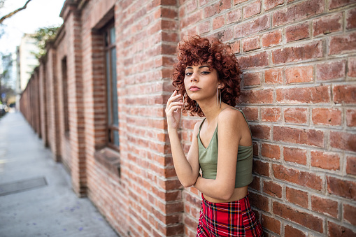 Portrait of young Latin woman with curly red hair standing in front of brick wall and  looking at camera seriously