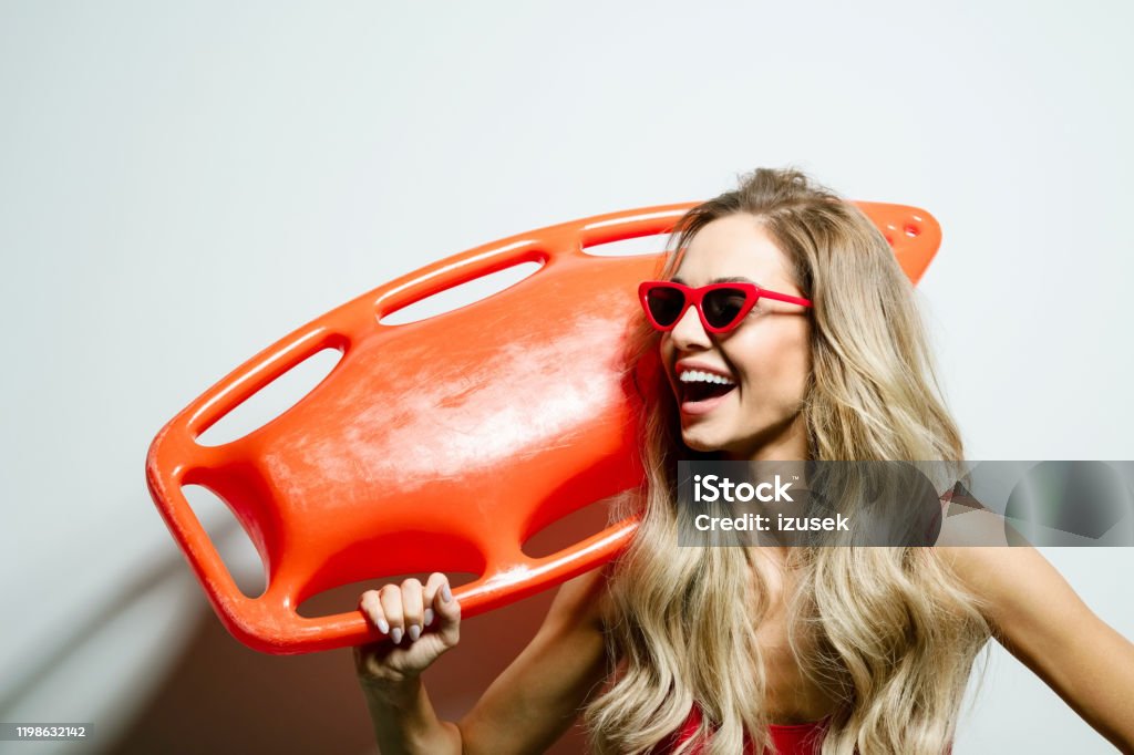 Summer portrait of young woman holding rescue buoy Portrait of happy beautiful long hair young woman wearing red swimsuit and sunglasses, holding orange rescue buoy on her shoulder. Studio shot on white background. 25-29 Years Stock Photo