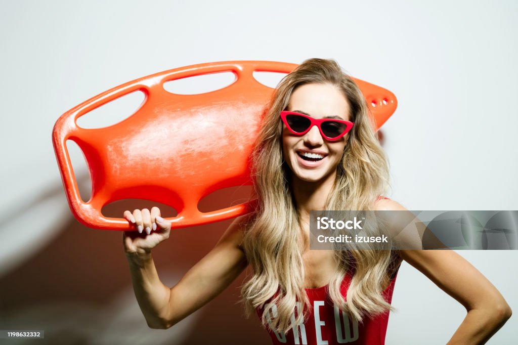 Summer portrait of young woman holding rescue buoy Portrait of happy beautiful long hair young woman wearing red swimsuit and sunglasses, holding orange rescue buoy on her shoulder. Studio shot on white background. Buoy Stock Photo