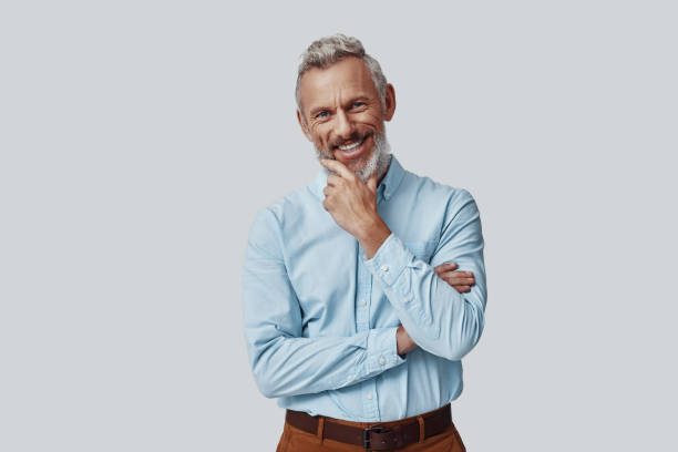 istock Happy mature man looking at camera and keeping hand on chin while standing against grey background 1198633071