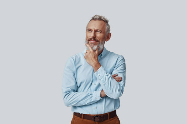 istock Thoughtful mature man looking at camera and keeping hand on chin while standing against grey background 1198633148