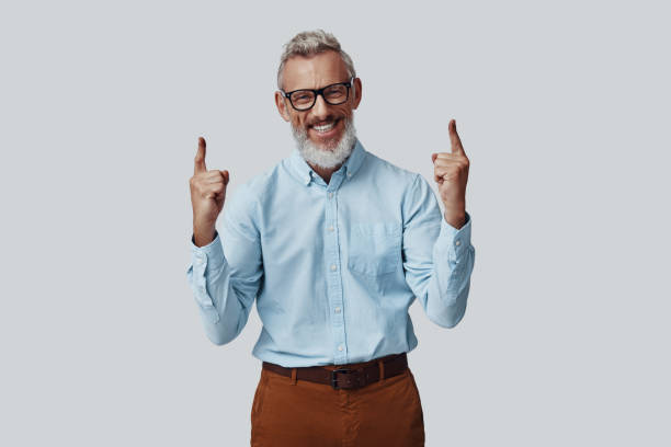 istock Cheerful mature man smiling and pointing copy space while standing against grey background 1198635765