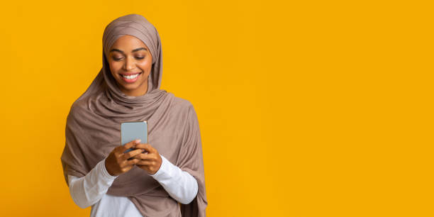 Cheerful Black Muslim Girl In Hijab With Smartphone Over Yellow Background Islamic Apps Concept. Cheerful Black Muslim Girl With Smartphone, Texting Or Browsing Internet Over Yellow Background, Panorama With Free Space arabian girl stock pictures, royalty-free photos & images