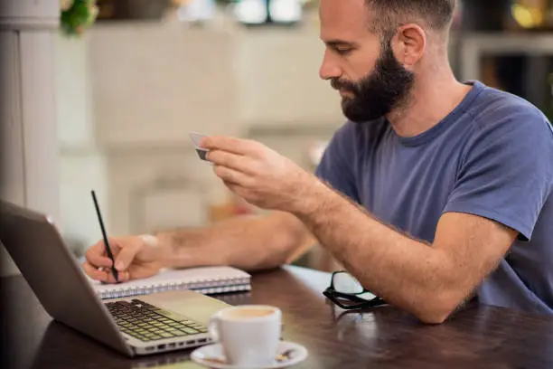 Photo of Handsome bearded caucasian man holding credit card and writing down outgoings while sitting in cafe.