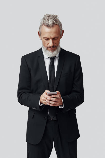istock Thoughtful mature man in full suit using smart phone while standing against grey background 1198638376