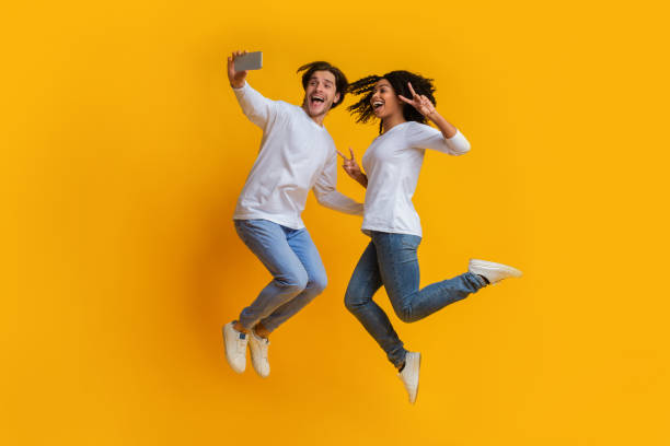 istock Millennial Interracial Couple Jumping And Taking Selfiem Posing Over Yellow Background 1198639103