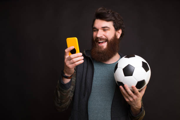 Happy man looking at smartphone and holding soccer ball, fan support team stock photo
