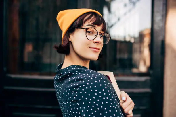 Portrait of smart young female student wearing yellow hat, transparent eyeglasses and green with white dots shirt with books in hands standing against the university.