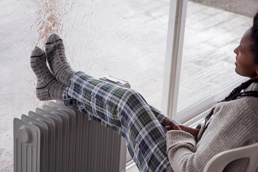 A relaxed woman comfortable sitting on the chair with legs on the radiator at home, looking through the large window in the winter rainy season. The woman wearing woolen socks and checkered pajama pants.