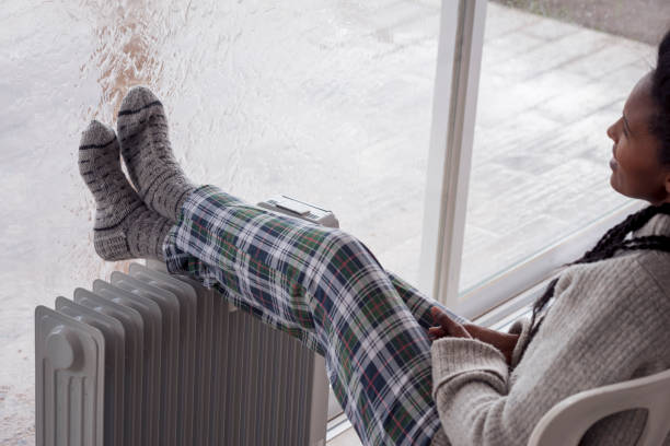 Woman heating legs in rainfall winter season, using an electric heater in cold weather. A relaxed woman comfortable sitting on the chair with legs on the radiator at home, looking through the large window in the winter rainy season. The woman wearing woolen socks and checkered pajama pants. electric heater photos stock pictures, royalty-free photos & images