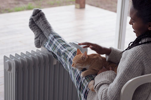 Woman stroking a ginger cat, comfortable sitting with legs on the radiator at home against the large window in winter rain season. The woman wearing woolen socks, using a heater in cold weather.