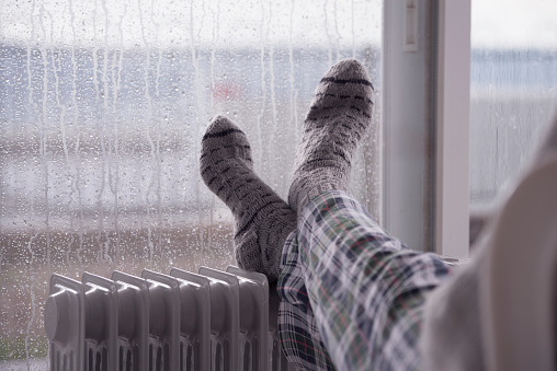 Woman wearing gray pair of woolen socks, warming cold feet in front of the heater, staying at home in the rain winter season.