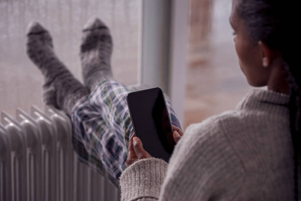 A woman heating up legs in front of an electric heater while using a smartphone. Over the shoulder view of woman using a mobile phone, warming cold feet in front of the heater, sitting against a wet window at home in rainy winter weather. Selective focus on a smartphone. woman hot shower stock pictures, royalty-free photos & images