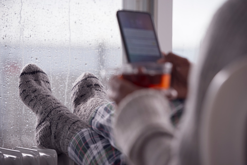 Woman using a smartphone, warming cold feet in front of the heater, sitting against a wet window at home in rainy winter weather. The woman drinking tea.