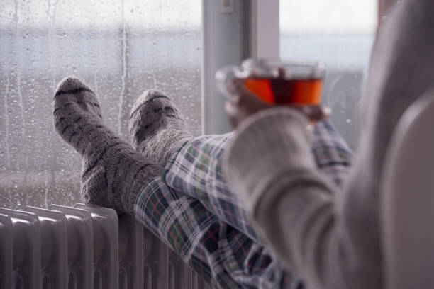 fermez-vous vers le haut chauffant des pieds femelles avec des chaussettes de laine sur le réchauffeur de radiateur. la femme buvant du thé chaud. - knitting residential structure glasses hot drink photos et images de collection