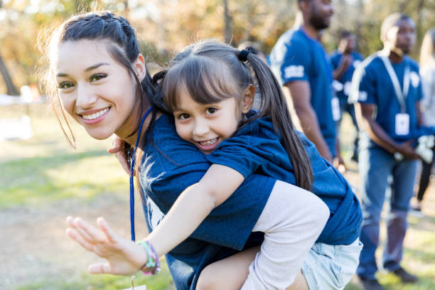 Sisters volunteering together Young girl rides piggyback on her big sister's back during a community cleanup event. They are smiling at the camera. social responsibility photos stock pictures, royalty-free photos & images