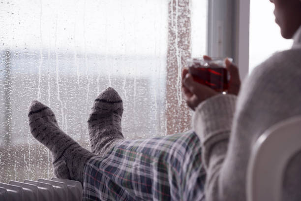 Over the shoulder image of a woman drinking tea at home in cold and wet weather. Over the shoulder view of a woman drinking hot tea, heating feet on the radiator heater, wearing woolen socks, sitting next to a window, staying at home in the rainy winter season. Selective focus on feet. woman hot shower stock pictures, royalty-free photos & images