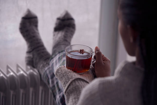 Over the shoulder image of a woman drinking tea at home in cold and wet weather. Over the shoulder view of a woman drinking hot tea, heating feet on the radiator heater, wearing woolen socks, sitting next to a window, staying at home in the rainy winter season. Selective focus on the cup of tea. cold temperature stock pictures, royalty-free photos & images