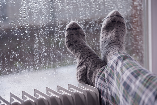 Woman's feet heating up on an oil radiator heater in front of a wet window. Selective focus on gray woolen socks. A woman staying at home in the rainy winter season.