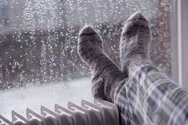 Woman's feet wearing gray woolen socks, warming cold feet on oil heater in the rainfall winter season. Woman's feet heating up on an oil radiator heater in front of a wet window. Selective focus on gray woolen socks. A woman staying at home in the rainy winter season. woman hot shower stock pictures, royalty-free photos & images