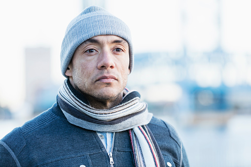 A mid adult Hispanic man in his 30s wearing warm clothing, a scarf and knit hat. He is standing in the city on the waterfront, bridge out of focus in the background. He is looking away with a serious expression.