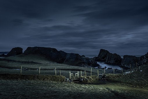 Wild rugged craggy irish coastline and bay at Ballintoy, Northern Ireland, a tourist attraction and where scenes from Game of Thrones were filmed, in early winter morning under dark cloudy moody sky and gate in foreground illuminated by light painting