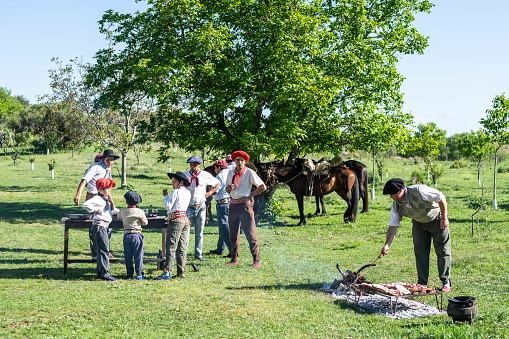 Argentine gaucho boys, teenagers, and adults enjoying springtime sunshine while meat is grilling for midday asado meal and horses rest under tree.