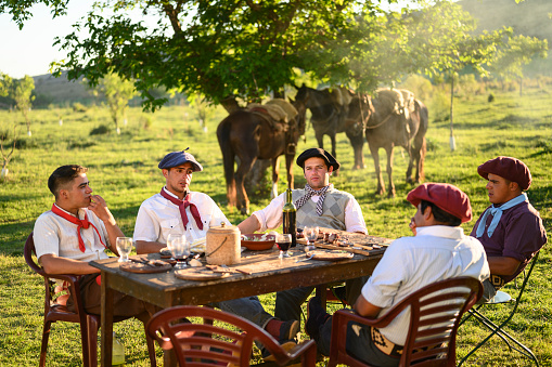 Five Argentine gauchos sitting at outdoor table relaxing after midday asado meal with wine on sunny spring day.