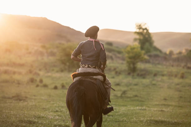 Young gaucho in traditional clothing riding in afternoon Rear view of Argentine 16 year old gaucho on horseback riding in grassland with sun setting over hills. gaucho stock pictures, royalty-free photos & images