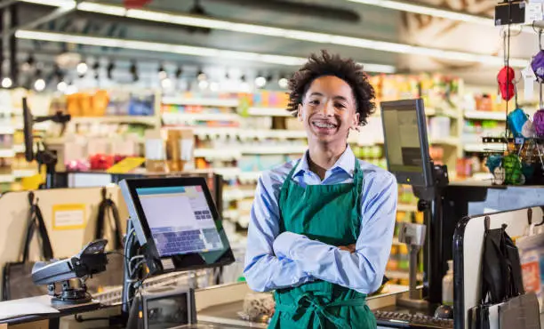 A mixed race African-American and Hispanic teenage boy working in a supermarket at the checkout counter. He is ready to scan groceries at the cash register. He is smiling at the camera.