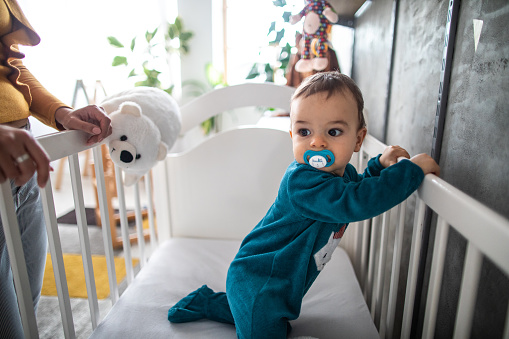 Cute baby boy in crib, who's wearing blue pajamas and has pacifier in his mouth trying to get up while mother watches him