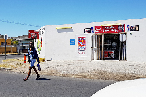 Cape Town, South Africa - December 16, 2019: A shopper carrying her purchases leaves a small convenience store in the lower-middle-class suburb of Grassy Park. Such shops, typically operated by recent immigrants from the Indian subcontinent or countries further north in Africa, have mushroomed across South Africa in recent decades.
