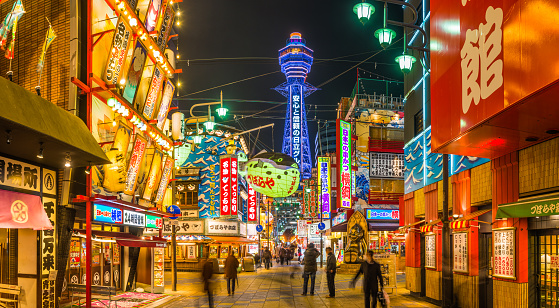 The iconic spire of Tsutenkaku Tower illuminated against deep blue dusk skies overlooking people strolling through the neon night streets of Shinsekai in the heart of Osaka, Japan's vibrant second city.