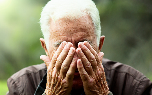 A senior man frowns, covering his face with his hands as he stands outdoors.