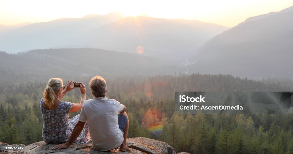 Mature couple relax on mountain ledge, look out to view She takes smart phone pic.The sun is rising ahead of them over the mountains Vacations Stock Photo