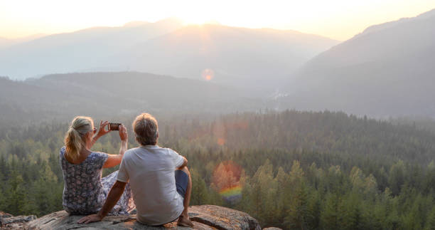 Mature couple relax on mountain ledge, look out to view She takes smart phone pic.The sun is rising ahead of them over the mountains mature men photos stock pictures, royalty-free photos & images