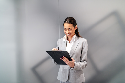 Young attractive Caucasian smiling businesswoman standing inside corporate firm and filling documents on clipboard.